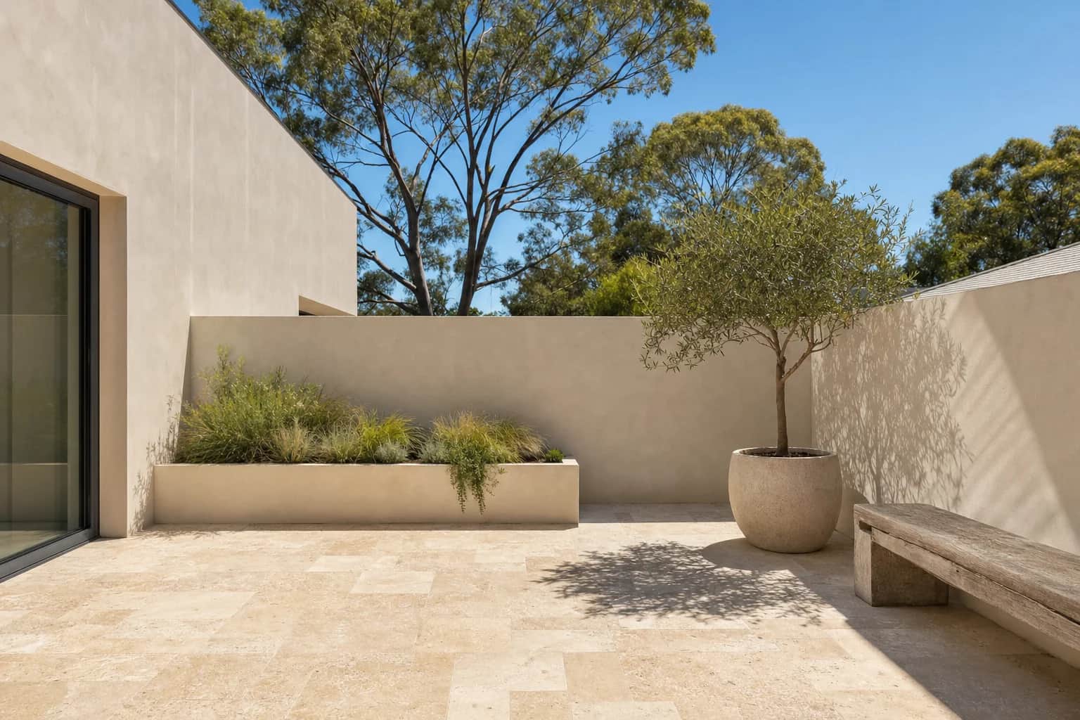 A small open courtyard paved in warm travertine with rendered walls and a potted olive tree in full sun