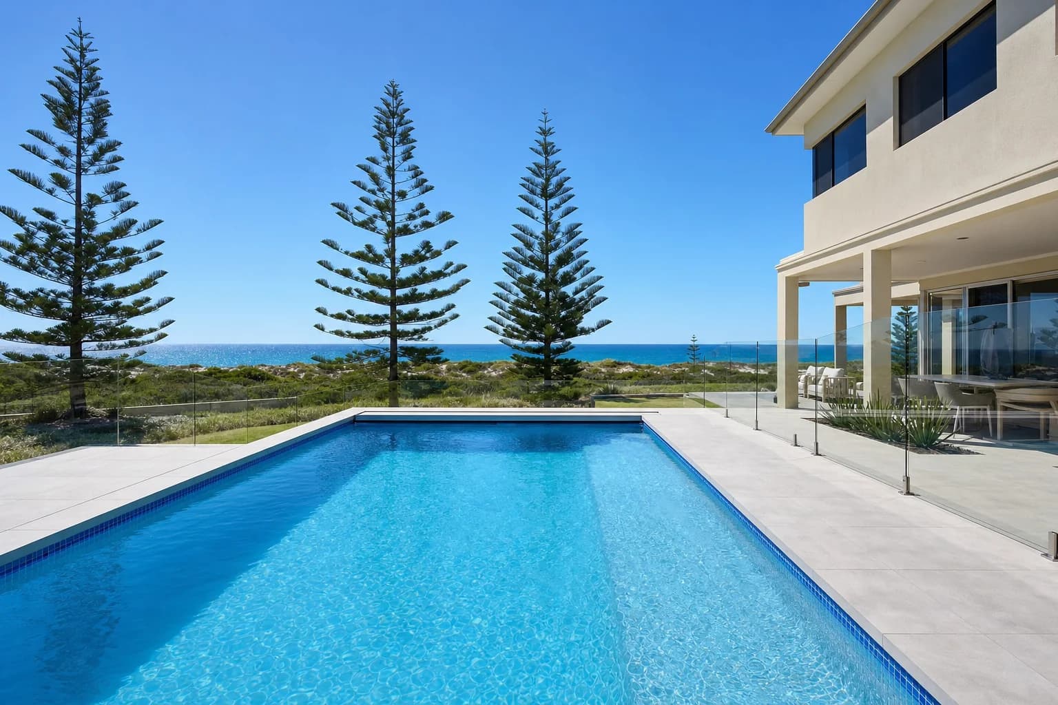 A pool surround in pale grey porcelain at a Hillarys coastal home, glass pool fence and the ocean past the dune