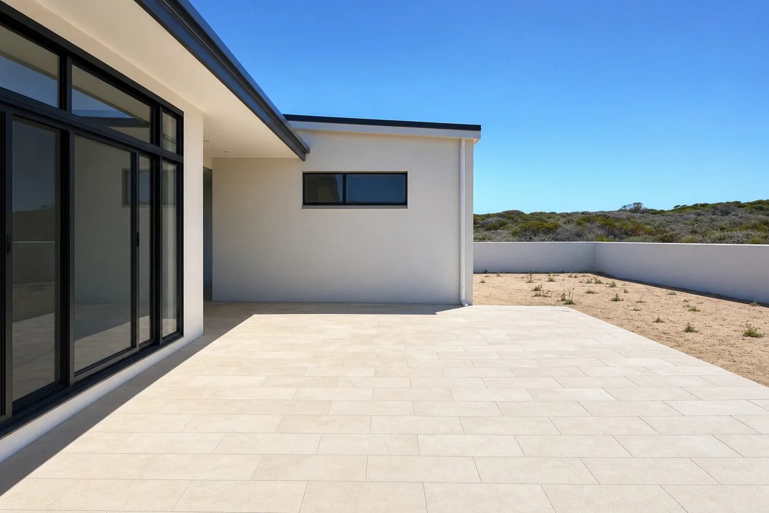 Cream limestone-look pavers laid across a new backyard in Two Rocks, bare sandy ground and a low boundary wall beyond