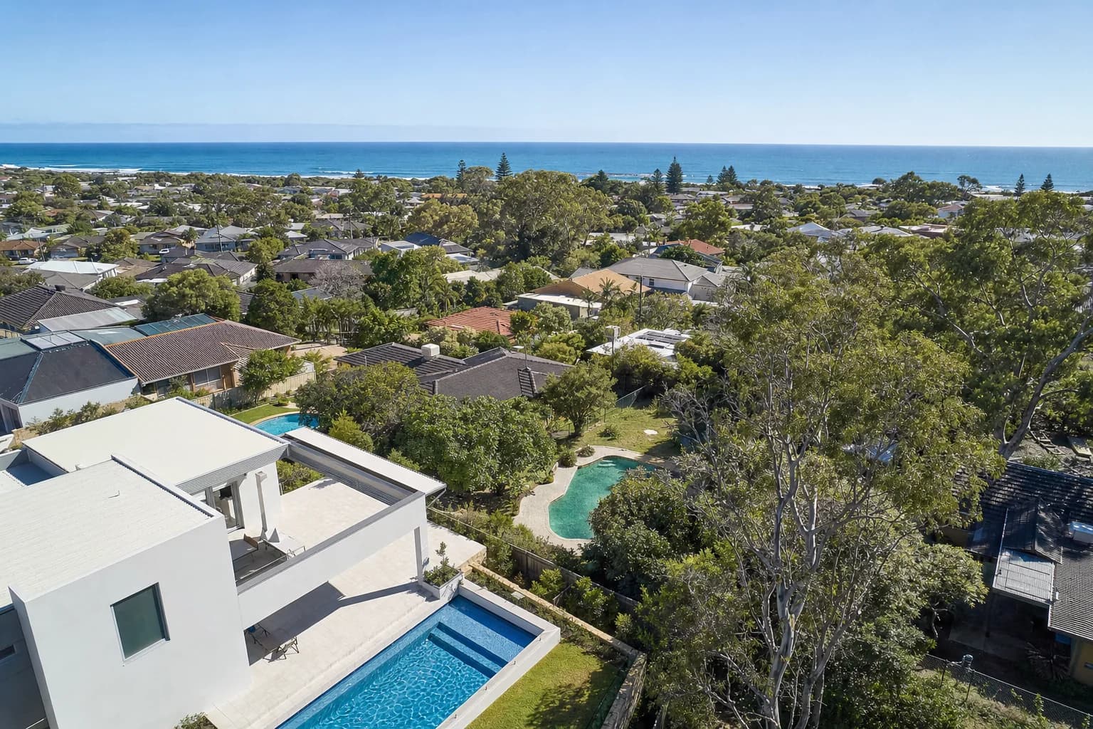A high wide view over a Perth coastal suburb of homes with swimming pools and paved backyards, the ocean along the horizon