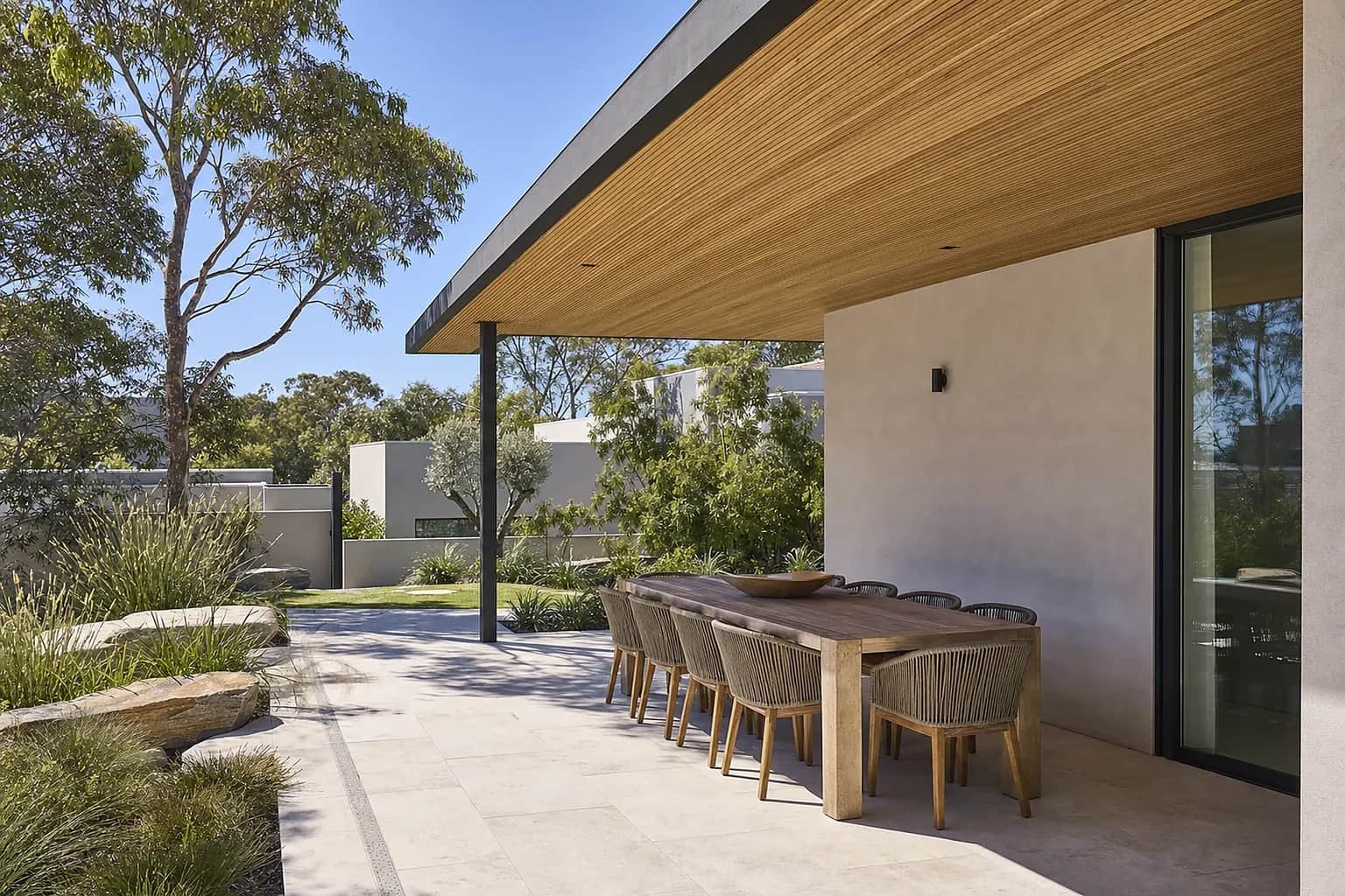 Pale porcelain paving under a covered alfresco with an outdoor dining table and a timber lined ceiling